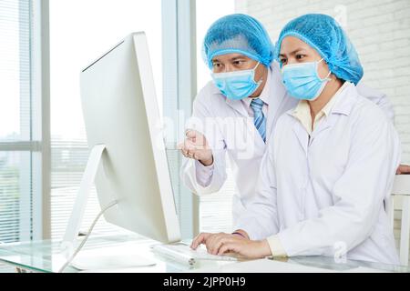Asiatische Ärztinnen und Ärzte in weißen Mänteln und medizinischen Masken versammelten sich in einem modernen Büro mit Panoramafenstern und diskutierten über Behandlungsmethoden ihres Patienten Stockfoto