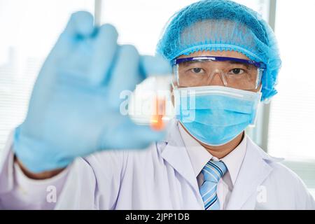 Asiatische Mikrobiologin mittleren Alters, die nach erfolgreichem Abschluss des Experiments einen weißen Mantel und eine Schutzbrille trägt und einen brandneuen Impfstoff in der Hand hält, Porträtaufnahme Stockfoto