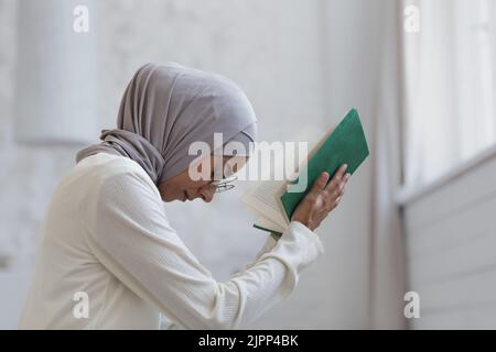 Eine junge, schöne muslimische Frau in einem Hijab betet zu Hause am Fenster Stockfoto