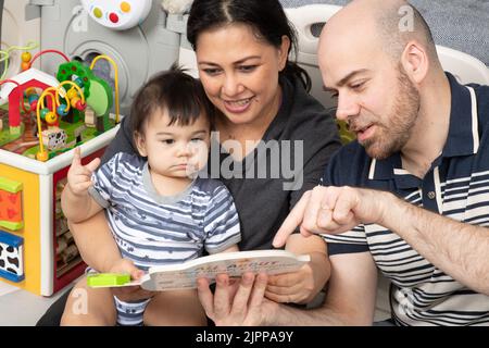 11 Monate alter Junge zu Hause von den Eltern vorgelesen, Vater mit Zeigefinger, um die Aufmerksamkeit auf etwas im Buch zu rufen, Baby hält Zeiger Figur Stockfoto