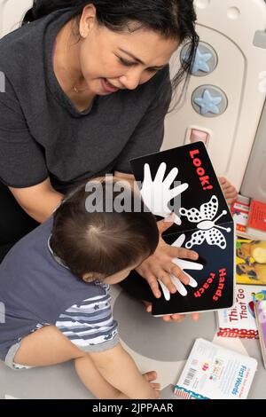 11 Monate alter Junge zu Hause mit Mutter, vorgelesen, interessiert, als sie ihre Hand über eine Abbildung einer Hand zeigt Stockfoto