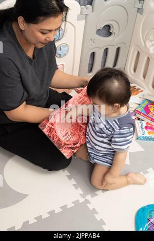 11 Monate alter Junge zu Hause mit Mutter aus dem Bordbuch mit Stoffeinsatz vorlesen, Baby berühren Bereich mit Finger Stockfoto