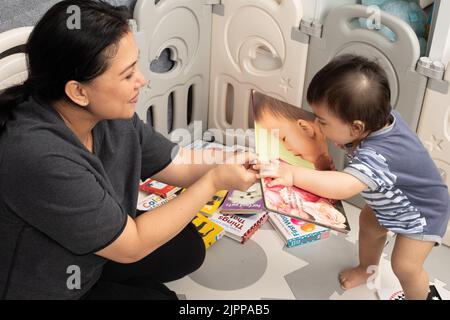 11 Monate alter Junge zu Hause, mit Mutter, vorgelesen, stehend, sich lehnend, um das Foto des Babys im Bordbuch zu küssen Stockfoto