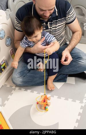 11 Monate alter Junge zu Hause mit Vater, der eine Schnur zieht, um ein Spielzeug auf Rädern näher zu bringen Stockfoto