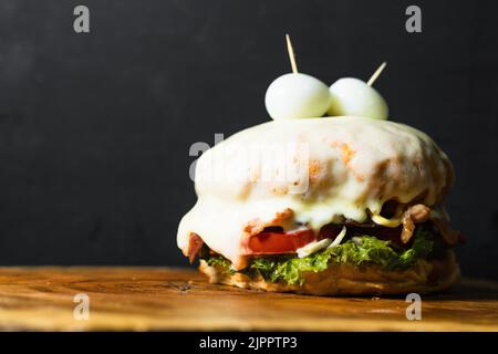 Kolumbianischer Hamburger in geschmolzenem Käse getaucht, serviert, mit zwei Eiern auf der Oberseite, die zwei Augen imitieren, auf einem Holztablett oder -Teller, auf schwarzem Hintergrund mit sp Stockfoto