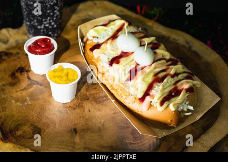 Draufsicht auf einen Hot Dog mit Saucen, Käse und zwei Wachteleiern auf einem Holztablett serviert und daneben zwei Plastikbecher mit Ketchup und Muste Stockfoto