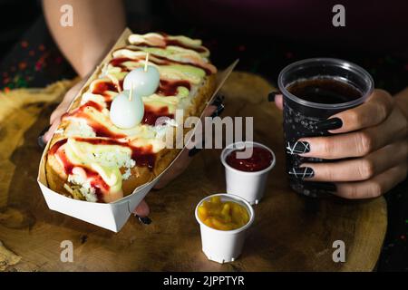 Mädchen hält in den Händen einen Hotdog mit Saucen, Käse und zwei Wachteleiern auf dunklem und hölzernen Hintergrund, daneben gibt es zwei Plastikbecher w Stockfoto