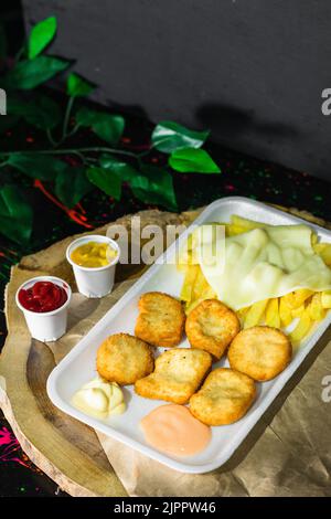 Nahaufnahme eines Tellers mit Hühnchen-Nuggets mit Kartoffeln und Mozzarella-Käse, serviert auf einem Holztablett mit zwei kleinen Gläsern mit Sauce. Schwarz Stockfoto