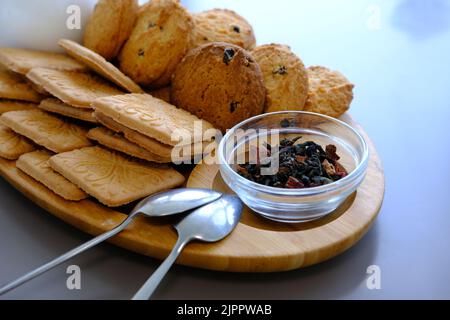 Getrocknete Teeblätter mit Teekannen und Keksen auf einem grauen Tisch mit Löffeln und Holzbrett. Stockfoto