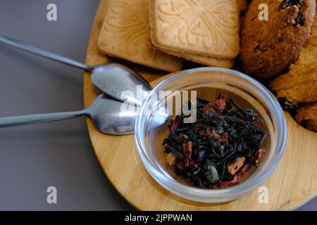 Getrocknete Teeblätter mit Teekannen und Keksen auf einem grauen Tisch mit Löffeln und Holzbrett. Stockfoto