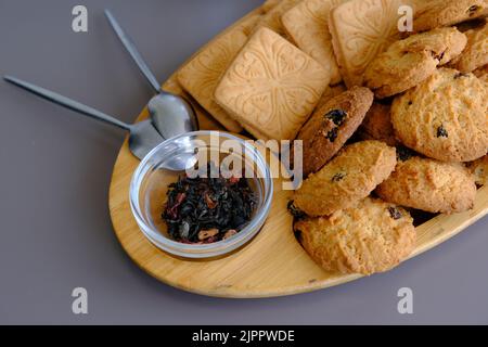 Getrocknete Teeblätter mit Teekannen und Keksen auf einem grauen Tisch mit Löffeln und Holzbrett. Stockfoto