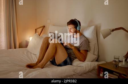 Laptop, Internet und entspannen Sie sich bei einem jungen Mann, der Musik, Videos oder einen Podcast online mit Technologie streamt und zu Hause auf seinem Bett sitzt Stockfoto