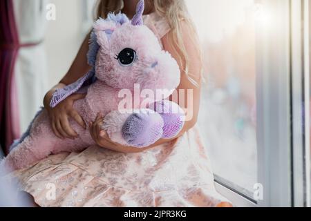 Kleines Mädchen hält ihr weiches Spielzeug Einhorn, während sie in der Nähe des Fensters sitzt Stockfoto