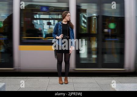 Sie fährt überall mit dem Zug. Eine attraktive junge Fotografin steht auf der U-Bahn mit einem vorbeifahrenden Zug im Hintergrund. Stockfoto