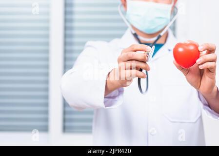 Nahaufnahme eines Arztes, der einen weißen Mantel trägt und sein Stethoskop zur Hand hält, um das rote Herz zu untersuchen und medizinische Geräte zu zeigen Stockfoto