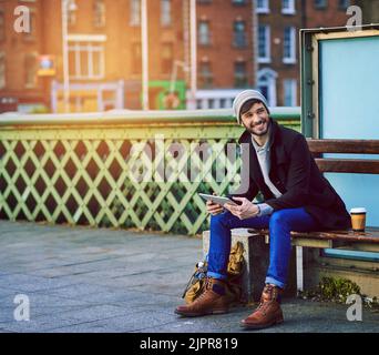 Ein junger Mann, der ein digitales Tablet benutzt, während er an einer Bushaltestelle in der Stadt wartet. Stockfoto