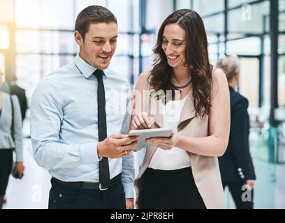 Zwei Köpfe sind besser als einer. Zwei junge Geschäftsleute, die in einem geschäftigen Büro an einem digitalen Tablet arbeiten. Stockfoto