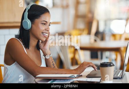 Glückliche Schülerin, die sich entspannt, studiert oder Online-Videos auf ihrem Laptop in einem Café mit kostenlosem Internet sucht. Junge Frau in einem Café hört zu Stockfoto