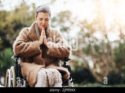 Alles, was ich brauche, ist ein Wunder. Eine ältere Frau hält ihre Hände im Gebet zusammen, während sie draußen in ihrem Rollstuhl sitzt. Stockfoto