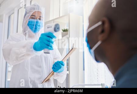 Gesundheitshelfer, der die Temperatur des männlichen covid Patienten auf Sicherheit vor Virus-Pandemie überprüft, trägt Schutzanzug. Arzt in einem Stockfoto