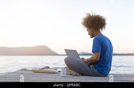 Junger digitaler Nomadenmann sitzt auf einem hölzernen Pier auf dem Meer und arbeitet bei Sonnenuntergang fernab im Internet - Reisen mit dem Computer - Online-Traumjob-Konzept Stockfoto