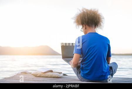 Junger Hipster-digitaler Nomadenmann sitzt auf einem hölzernen Pier am Meer und arbeitet im Internet bei Sonnenuntergang aus der Ferne - Reisen mit dem Computer - Online-Traumjob Co Stockfoto