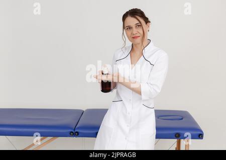 Portrait der wunderschönen Masseuse mittleren Alters tragen weißes Kleid, stehen in der Nähe der blauen Couch, halten Flasche mit Massageöl. Stockfoto