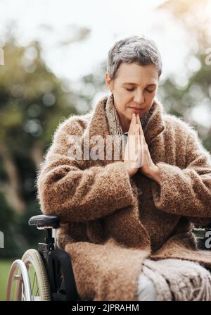 Eine ältere Frau hält ihre Hände im Gebet zusammen, während sie draußen in ihrem Rollstuhl sitzt. Stockfoto