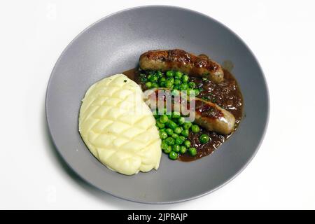 Pürieren und Maische, gegrillte Schweinewurst mit Kartoffelpüree, brauner Zwiebelsoße und gedämpften grünen Erbsen Stockfoto