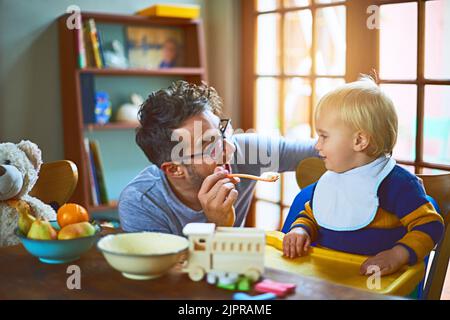 Papa macht immer Spaß beim Essen. Ein alleinerziehender Vater füttert seinen Sohn zu Hause. Stockfoto