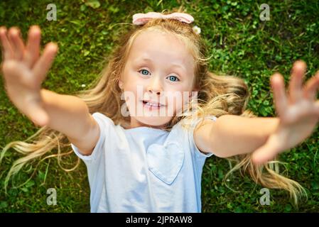 Nimm mich ab. Porträt eines fröhlichen kleinen Mädchens, das auf Gras liegt und ihre Arme ausstreckt, während es draußen auf die Kamera schaut. Stockfoto