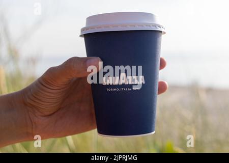 Eine junge Frau hält an einem sonnigen Morgen am Strand eine Tasse Lavazza-Kaffee zum Einwegartikel. Kaffee zum Mitnehmen am Meer. Turin, Italien Stockfoto
