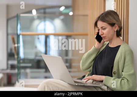 Seriöse Geschäftsfrau diskutiert über Projektdetails per Handy, während sie ferngesteuert am Laptop arbeitet Stockfoto