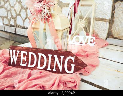 Schöne Hochzeitsdekoration auf steinigen Treppen mit Holzpfeilhochzeit. Große Kerzenlaternen mit rosa Blüten und Bändern auf staubigen rosa Stoff outsid Stockfoto