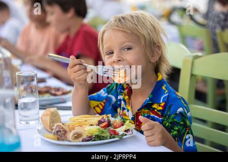 Süßes Kind, das im Sommer im Restaurant im Freien sitzt und Meeresfrüchte, Garnelen, Calamari, Tintenfisch und pommes frites isst Stockfoto