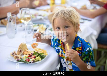 Süßes Kind, das im Sommer im Restaurant im Freien sitzt und Meeresfrüchte, Garnelen, Calamari, Tintenfisch und pommes frites isst Stockfoto