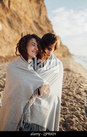 Junge, fröhliche Frau und Mann, die bei Sonnenaufgang am Meer in ein Karloid gehüllt stehen Stockfoto