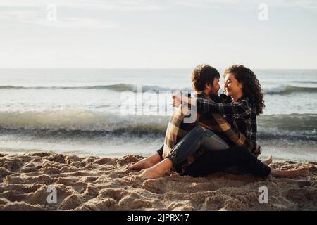 Junges Paar, lockige Frau mit brünetem Mann verlieben sich beim Sonnenaufgang am Meer sitzend und umarmt Stockfoto