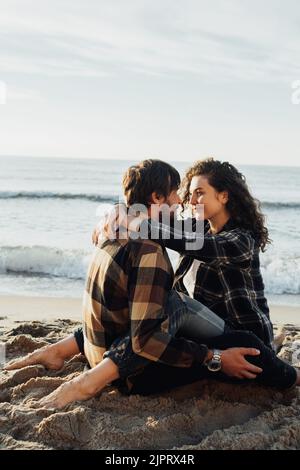 Junge Frau und Mann umarmen sich, während sie bei Sonnenaufgang zusammen am Meer sitzen und Meereswellen im Hintergrund beobachten Stockfoto