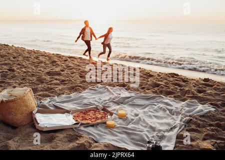 Konzentrieren Sie sich auf Picknick-Set mit Pizza und Saft, glückliches Paar, Frau und Mann an den Händen halten und laufen entlang der Küste zusammen im Hintergrund Stockfoto