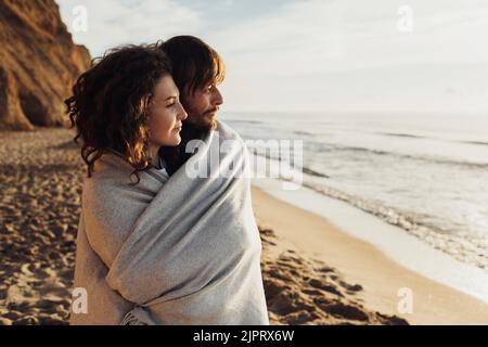Ein junges erwachsenes Paar, das in ein Karabinet gehüllt an der Küste steht und bei Sonnenaufgang auf die Meereswellen blickt Stockfoto