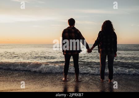 Junges Paar trifft Sonnenaufgang am Meer, Mann und Frau halten sich an Händen, die im Wasser stehen Stockfoto