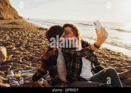 Glückliches junges erwachsenes Paar, das sich verliebt, eine lockige Frau, die zusammen mit ihrem Freund Selfie macht, während sie im Morgengrauen am Meer sitzt Stockfoto