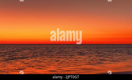 Leuchtender orange-roter Himmel, der sich nach Sonnenuntergang in einem ruhigen Meer widerspiegelt. Minimales Landschaftsfoto mit Platz für Text Stockfoto