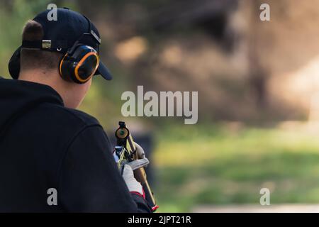 Rückansicht eines Mannes mit schwarzem Hoodie und Kappe, der Sicherheitskopfhörer trägt, um die Waffe aufzuladen. Waffenausbildung. Brennweite. Außenaufnahme aus Horiozntal. Hochwertige Fotos Stockfoto
