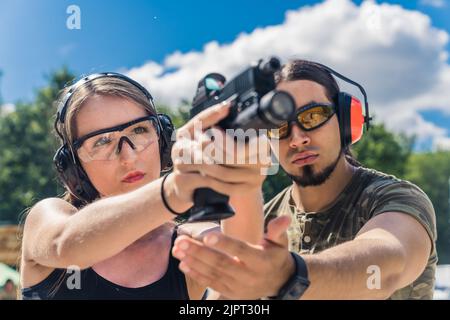 Weißer bärtiger Mann im Camo-T-Shirt, der kaukasische Frau anweist, wie man mit der Handfeuerwaffe anvisiert. Sicherheitsausrüstung. Schusswaffenausbildung. Zielgerichtete Übung. Horizontale Aufnahme . Hochwertige Fotos Stockfoto