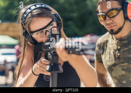 Kaukasische Frau mit Sicherheitskopfhörern und Schutzbrille, die auf eine Maschinenpistole zielte, beobachtet von einem bärtigen Mann im Tarnhemd. Schusswaffentraining im Feuerwehrbereich. Horizontale Aufnahme. Hochwertige Fotos Stockfoto