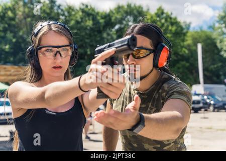 Weißer bärtiger Mann, der kaukasische Frau anweist, wie man mit einem Handfeuerwaffen zielt. Sicherheitsausrüstung. Schusswaffentraining auf dem Schießstand. Horizontale Aufnahme im Freien. Hochwertige Fotos Stockfoto