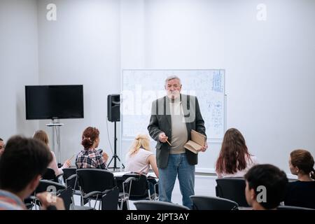 Ein männlicher Lehrer in einer Jacke hält an der Universität einen Vortrag über Mathematik, er steht an der weißen Tafel, Studenten sitzen in einem großen Au Stockfoto