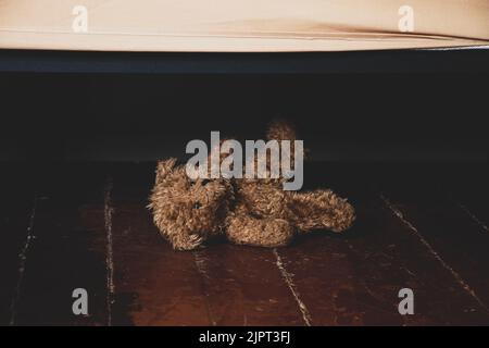 Neben dem Bett auf dem Boden im Schlafzimmer des Hauses liegt ein Teddybär, ein Teddybär unter dem Bett auf dem Boden im Dunkeln Stockfoto